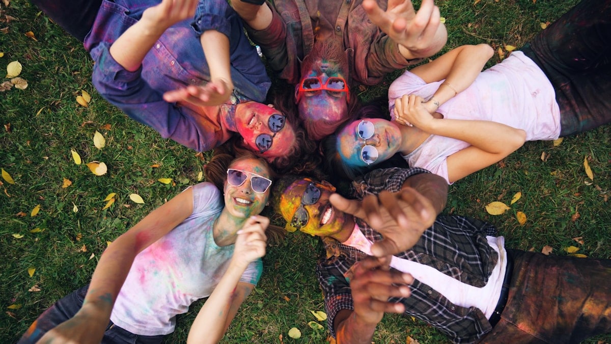 Joyful girls and guys are lying on grass in park, their faces and clothing are covered with bright multicolor paint, people are moving hands and smiling looking at camera.